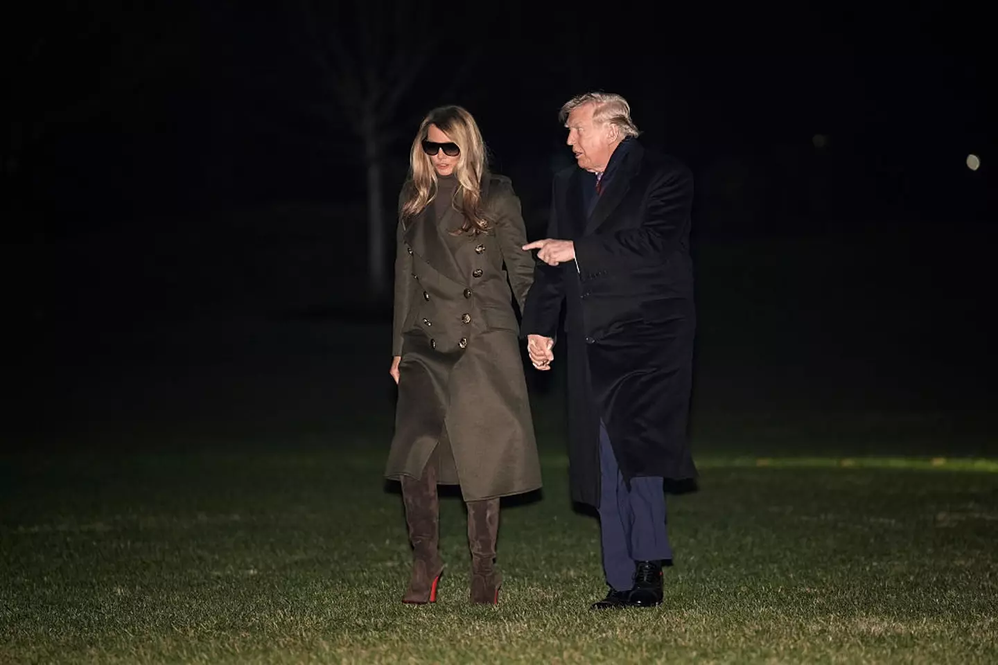 A close-up of Melania Trump wearing a dark coat and large sunglasses at night, with Donald Trump waving beside her.