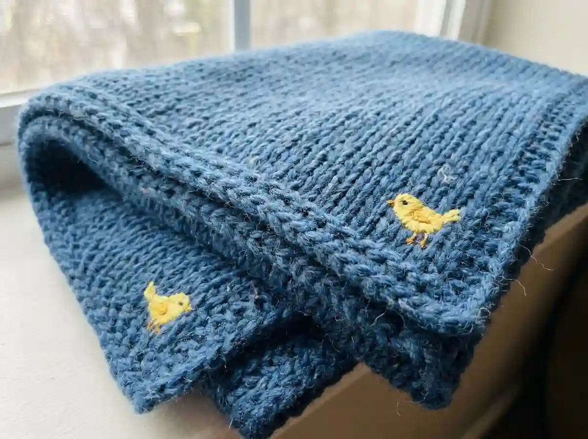 A folded blue wool baby blanket with tiny yellow birds stitched in the corners