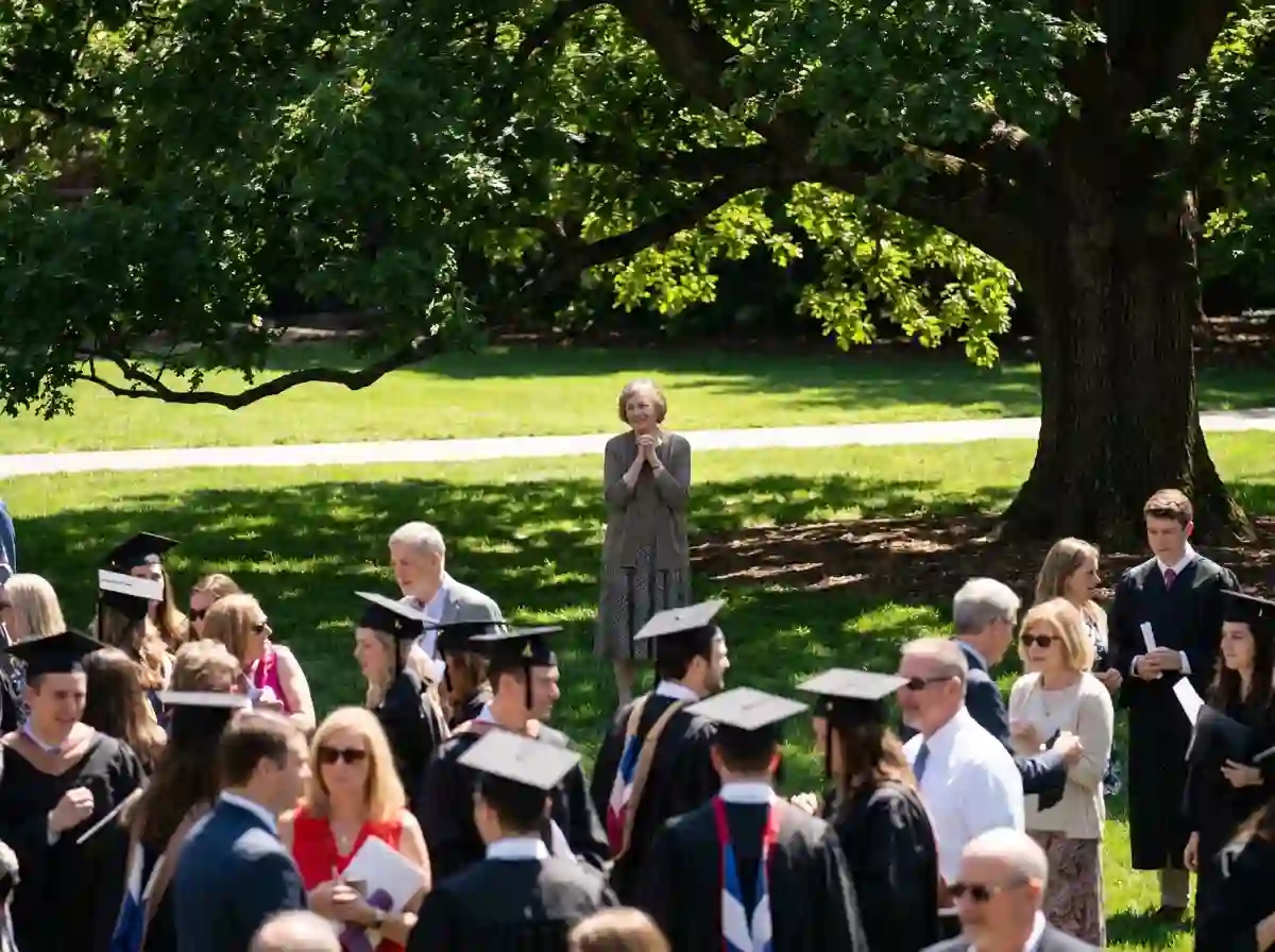 An older woman watching a graduation ceremony from a distance under a tree