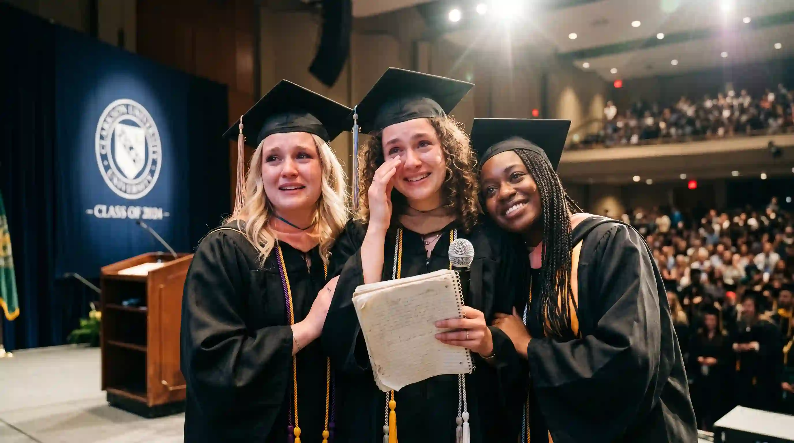 Three sisters on a graduation stage reading from an old notebook.
