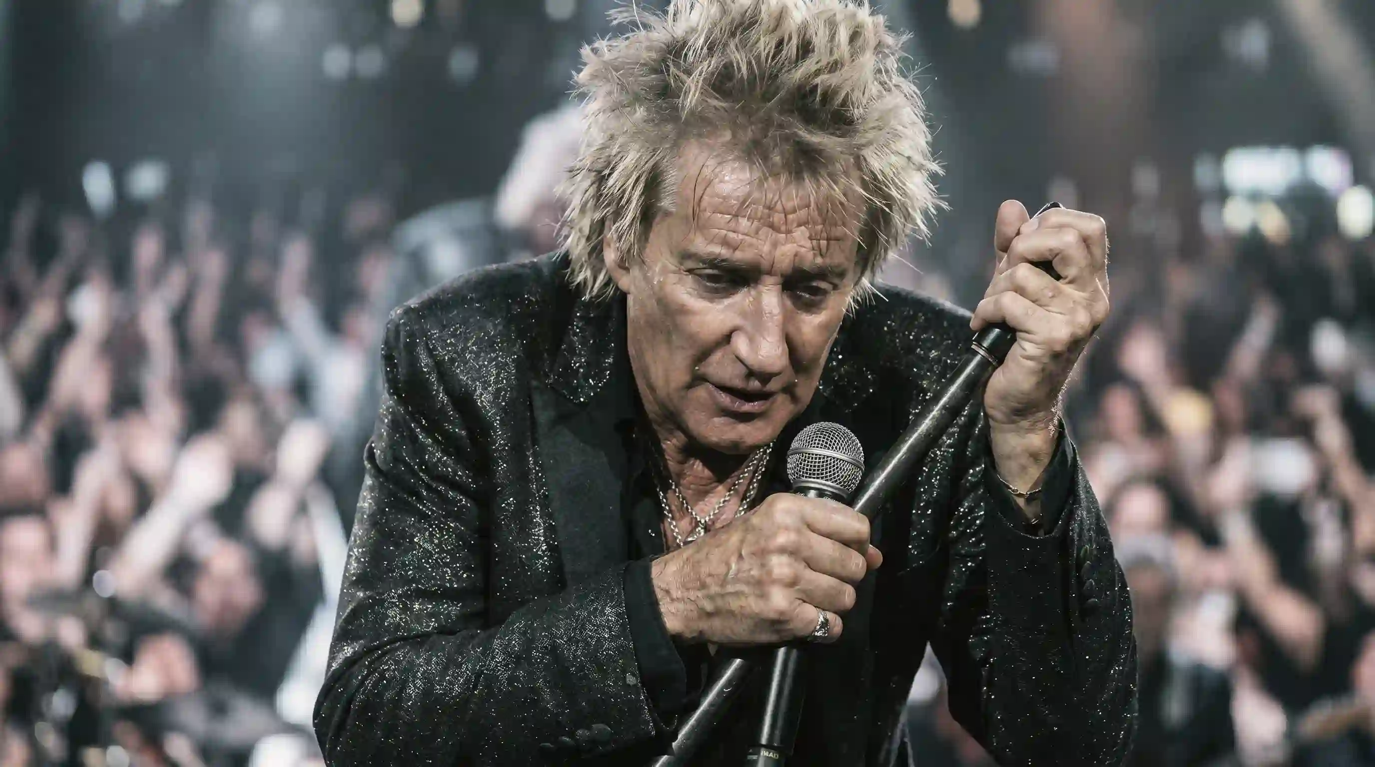 A zoomed-in dramatic photo of Rod Stewart, looking pale, gripping the microphone stand with both hands for support, looking distressed.
