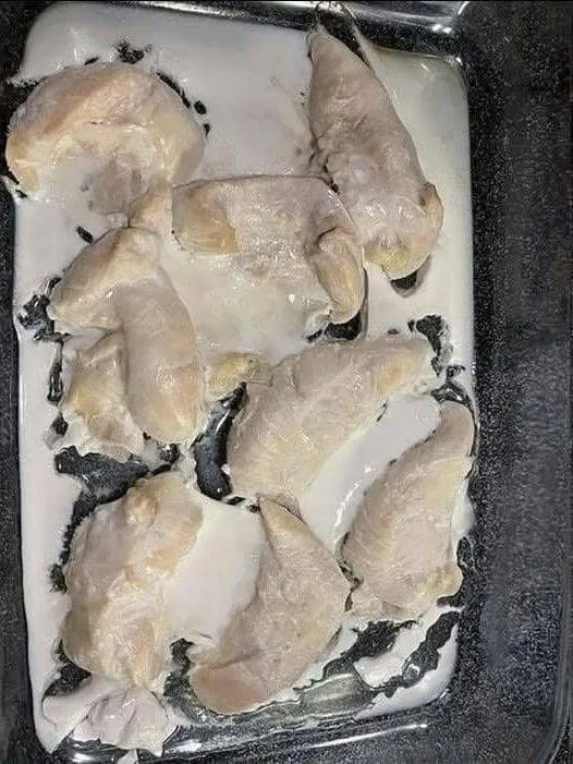 Fresh raw chicken breasts on a rustic wooden cutting board being patted dry with a paper towel, surrounded by fresh herbs.