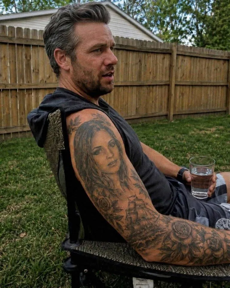 A realistic black and grey tattoo of a sad young woman's face on a man's shoulder.
